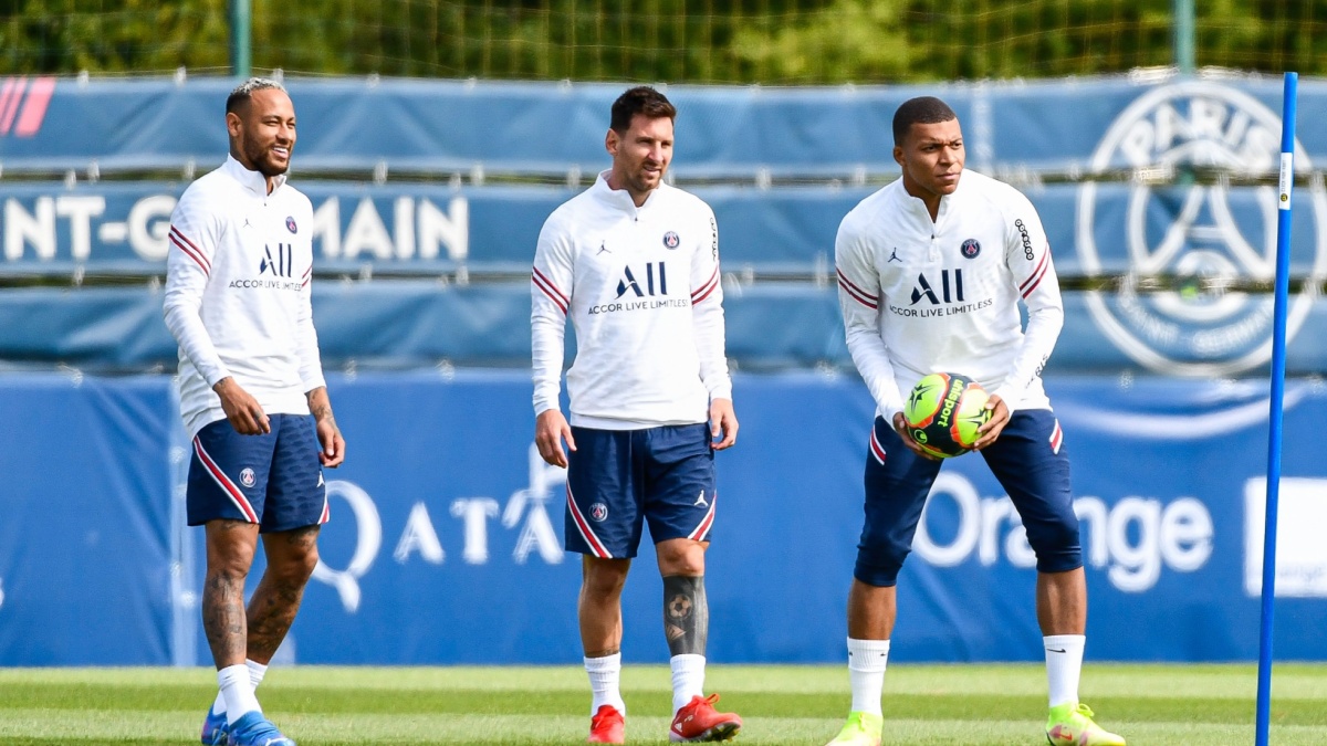 Neymar, Messi, and Mbappe in PSG training gear are on a practice field. One is holding a ball, while the others smile. A PSG banner is in the background.